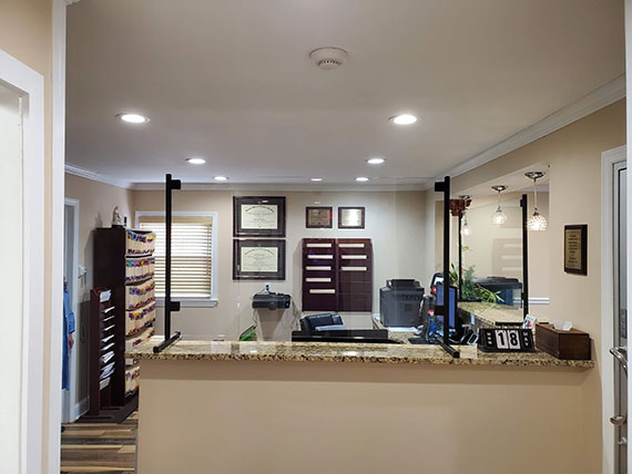 The image shows an interior view of a professional office space, featuring a reception desk with a computer monitor and keyboard, a countertop with a glass display case containing items, a wooden cabinet with drawers, a black and white checkered floor pattern, and a wall-mounted clock.