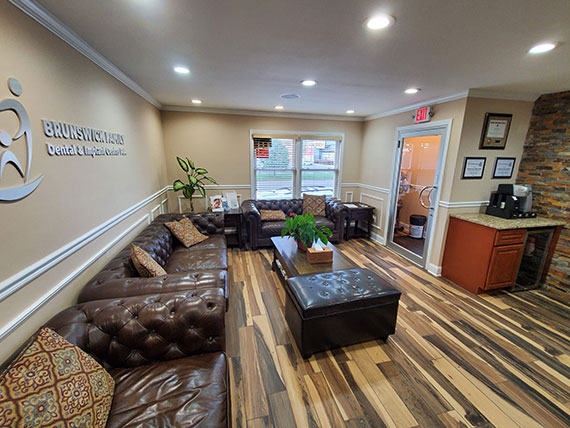 The image shows an interior view of a waiting room with a modern design, featuring a reception desk, seating area with leather sofas and ottomans, a coffee table, and a hardwood floor.