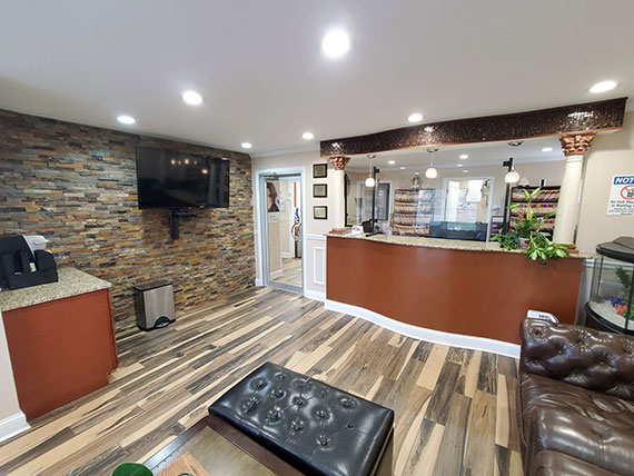 The image shows an interior view of a room with a counter, a television mounted on the wall, and a reception desk. The walls are adorned with brickwork and the flooring has wood-like planks. There is a seating area with a couch and a coffee table.