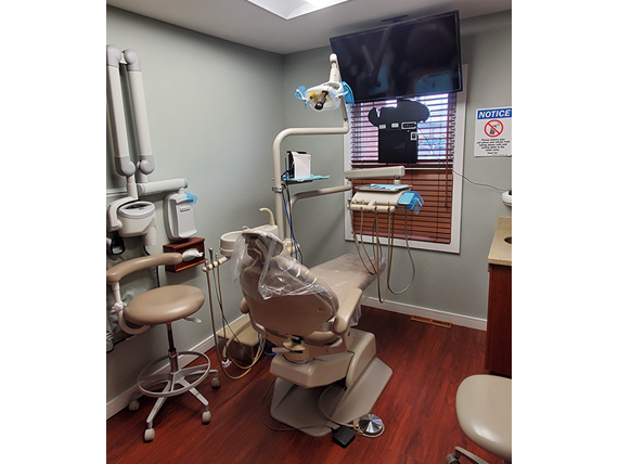 The image depicts a dental office interior, featuring a dental chair with a patient s headrest and a dental light, along with various dental equipment and a monitor displaying what appears to be an X-ray or digital imaging.