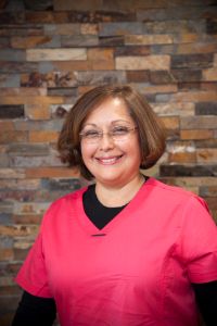 The image is a portrait of a woman wearing a red shirt and black pants, standing in front of a brick wall.