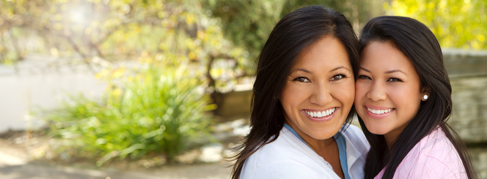 Two smiling individuals, a woman and a girl, embracing in an outdoor setting with greenery.