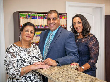 The image shows three individuals posing for a photo in an office setting. There is a man standing behind a counter, flanked by two women  all are smiling and appear to be engaged in a professional environment.