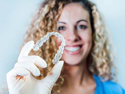 The image shows a person holding up a transparent dental retainer, smiling and wearing a white coat.