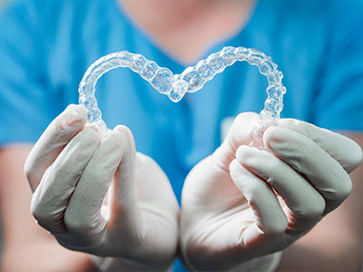 A dental professional in a blue uniform is holding a transparent heart-shaped object, which appears to be a clear aligner or orthodontic appliance, against their chest.