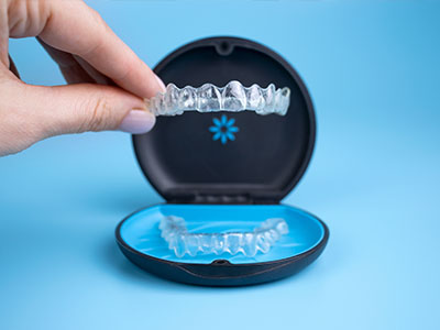 The image shows a person s hand holding an open box containing a set of clear, transparent dental aligners, with one aligner partially inserted into the mouth.