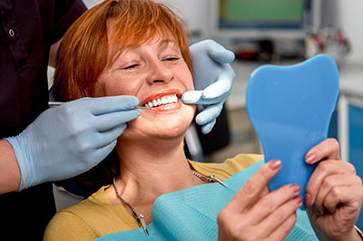 A woman sitting in a dental chair, smiling as she looks at her teeth, with a dental professional adjusting her mouthpiece.