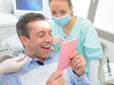 The image depicts a man sitting in a dental chair, holding up a pink card with a smile on his face, while a dental professional is standing behind him, smiling and looking at the card.