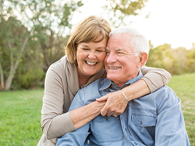 A man and woman in a loving embrace, both smiling, with the man wearing glasses and a blue shirt. They are outdoors during daylight, under a clear sky.