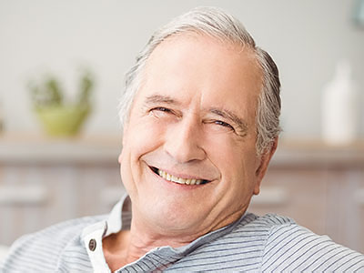 The image shows an elderly man with gray hair, wearing a blue shirt and smiling broadly. He is seated in what appears to be his home, with a warm and inviting atmosphere.